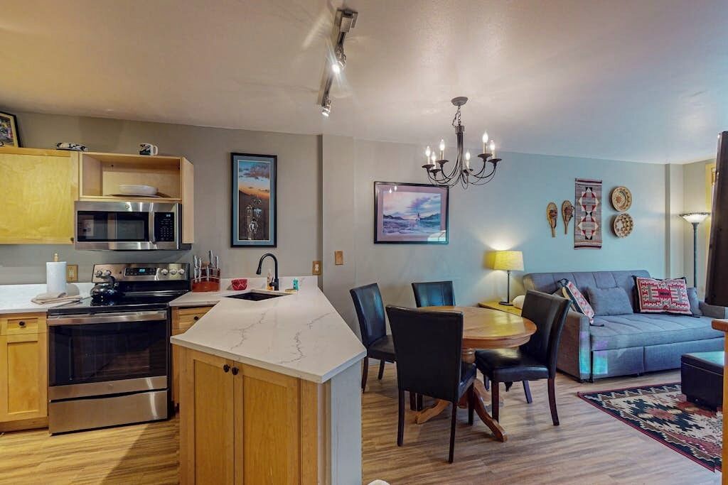 A modern kitchen with a built-in microwave, oven, and sink. There is a dining area with a table and chairs, and a living room with a sofa and a chandelier.