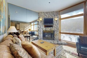 Una sala de estar con chimenea de piedra, un televisor montado encima y un ventanal con vistas a un paisaje nevado.