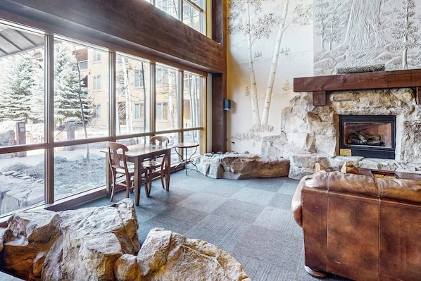Ein gemütliches Interieur mit Kamin aus Stein, einer Ledersofa, einem Esstisch mit Stühlen und großen Fenstern mit Blick nach draußen.