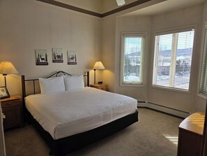 A bedroom with a bed, two bedside lamps, a nightstand, a framed picture, and a window with a view. - Comfortable Condo w/ Shared Pool, Gym, Hot Tub, Winter Shuttle, & Ski Lockers (Steamboat Springs)