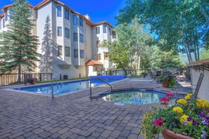 A swimming pool area with a brick patio, a building with multiple windows, and surrounding trees. - Comfortable Condo w/ Shared Pool, Gym, Hot Tub, Winter Shuttle, & Ski Lockers (Steamboat Springs)