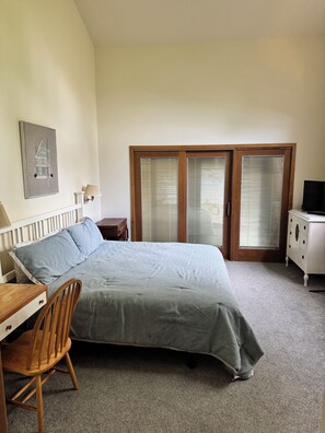 Ein Schlafzimmer mit einem Bett, einem Schreibtisch, einem Stuhl, einer Kommode, einem Fernseher und einem Fenster mit Jalousien.