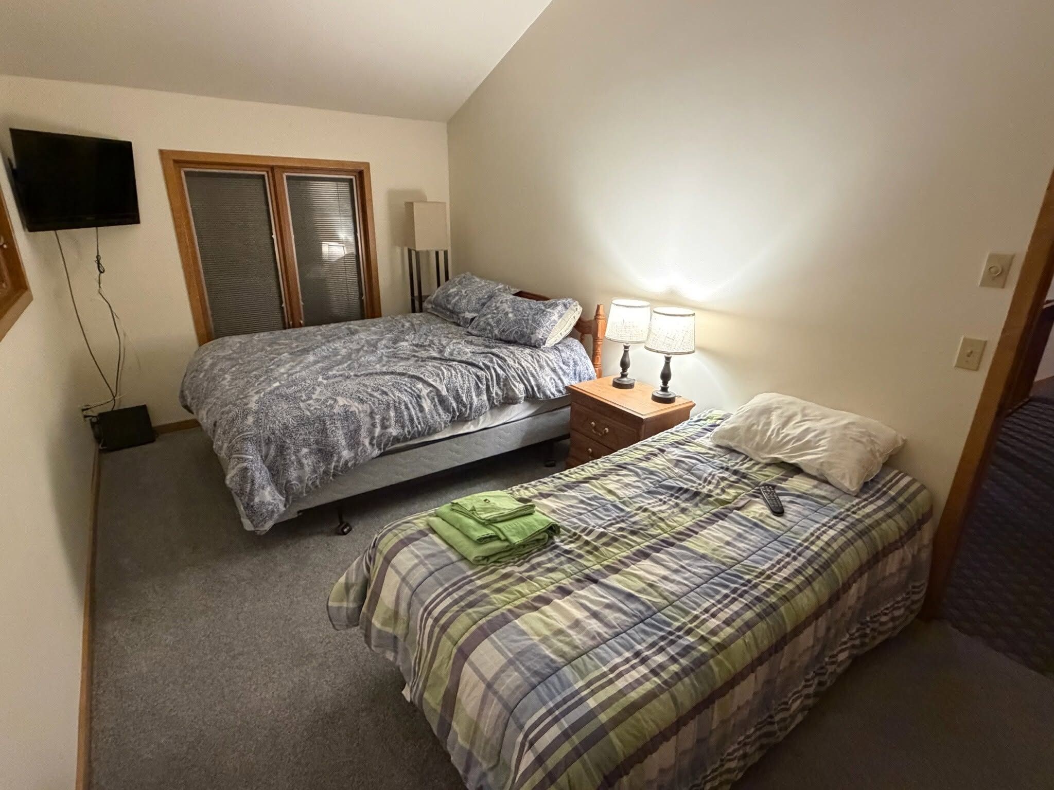 A bedroom with two beds, a TV mounted on the wall, and a bedside table with lamps.