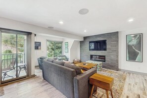 Ein modernes Wohnzimmer mit einem Ecksofa, einem an einer Steinwand montierten Fernseher und einem Essbereich mit Tisch und Stühlen.