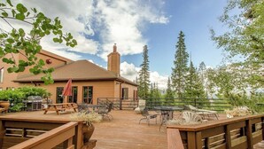 Una terraza de madera con mobiliario exterior, una parrilla y una edificación con chimenea.