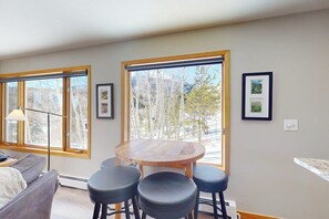 Ein Zimmer mit einem runden Holztisch, vier schwarzen Hocker, einem grauen Sofa und großen Fenstern mit Blick auf Bäume und Berge.