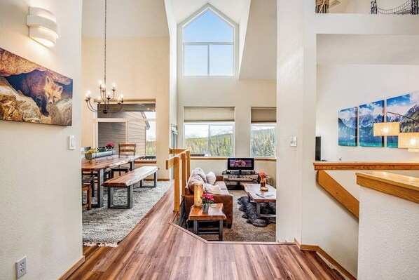 A spacious interior with a high ceiling, wooden flooring, a dining area, a seating area with a fireplace, and large windows offering views. - Home with hot tub, gas fireplace, and garage (Silverthorne)