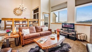 A spacious living room with a brown leather sofa, a wooden coffee table, a TV stand, and a large window offering a view of the outdoors. - Home with hot tub, gas fireplace, and garage (Silverthorne)
