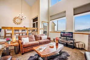 A spacious living room with a brown leather sofa, a wooden coffee table, a TV stand, and a large window offering a view of the outdoors. - Home with hot tub, gas fireplace, and garage (Silverthorne)