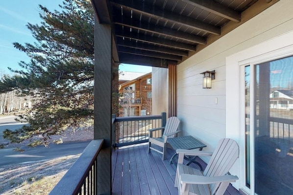 Eine überdachte Veranda mit zwei Adirondack-Stühlen, einem Tisch und Blick auf ein Gebäude und Bäume.