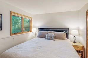Ein Schlafzimmer mit einem großen Bett, zwei Nachttischlampen, einem Fenster mit Blick auf Bäume und einem Holznachttisch.