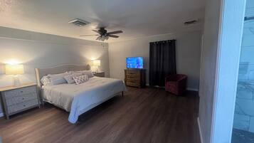 Een slaapkamer met een bed, twee nachtkastjes met lampen, een plafondventilator, een televisie op een houten dressoir, een rode stoel en een raam met gordijnen.