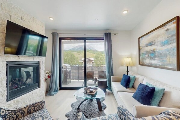 A living room with a fireplace, a sofa, a coffee table, and a TV mounted on the wall. - Cozy condo w/ full kitchen, fast wifi, balcony & convenient location (Silverthorne)