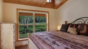 A bedroom with a large window, a bed with patterned bedding, a wooden headboard, and a nightstand.