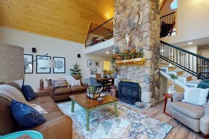 A spacious living area with a stone fireplace, a wooden ceiling, and a staircase leading to an upper level.