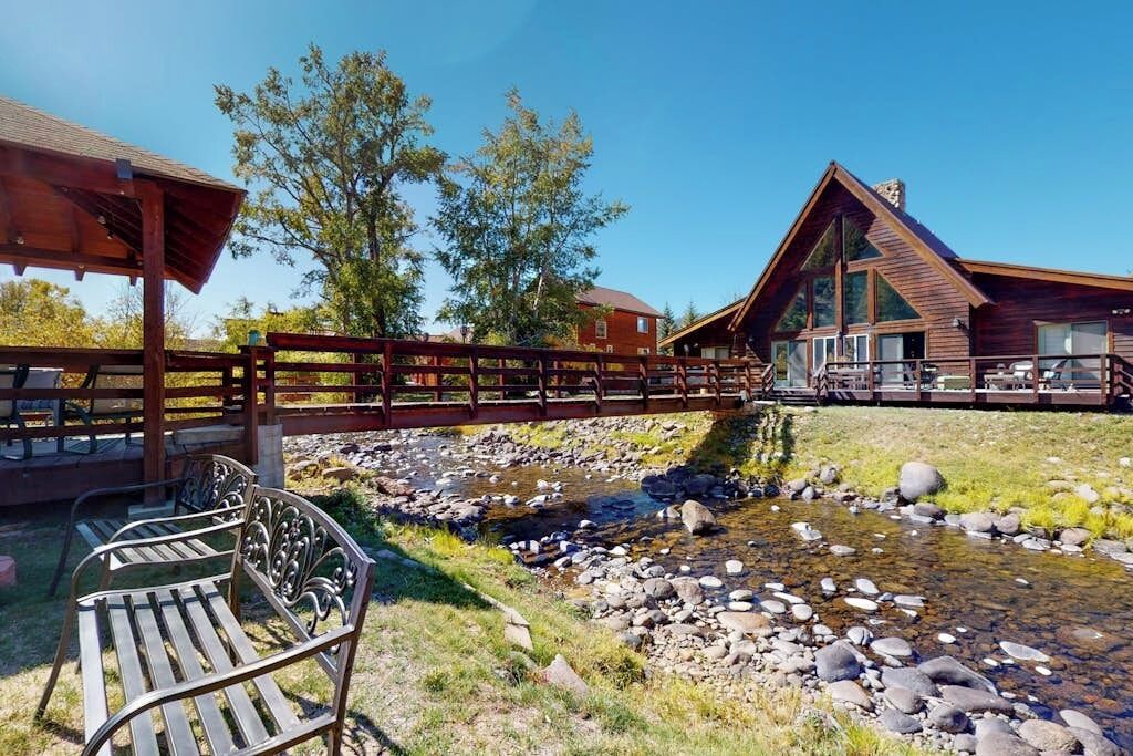 Eine Holzbrücke über einen Bach, eine hölzerne Bank und eine Lodge mit großen Fenstern.