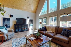 A spacious living room with a high ceiling, large windows, a television, a sofa, a coffee table, and a fireplace.