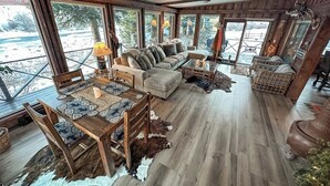 Un intérieur de chalet chaleureux avec un coin repas, un canapé et de grandes fenêtres donnant sur un paysage enneigé.