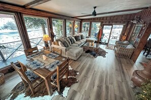 Ein gemütliches Blockhauss-Interieur mit Essbereich, Sofa und großen Fenstern mit Blick auf eine verschneite Landschaft.