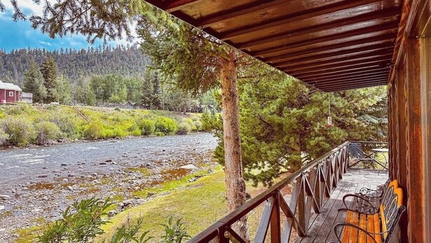 Une terrasse en bois avec un banc donnant sur une rivière, entourée d’arbres et d’un bâtiment situé au loin.
