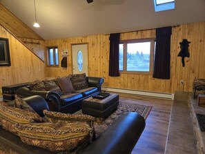 Ein Holzblockhaus-Interieur mit Ledersofa, gemusterten Kissen, Teppich, Kamin und großem Fenster mit Vorhängen.
