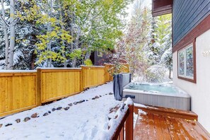 Eine Holzterrasse mit Whirlpool, umgeben von schneebedeckten Bäumen und einem Holzzaun.