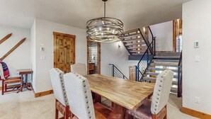 Un comedor con una mesa y sillas de madera, una lámpara colgante, una escalera y una puerta de madera.