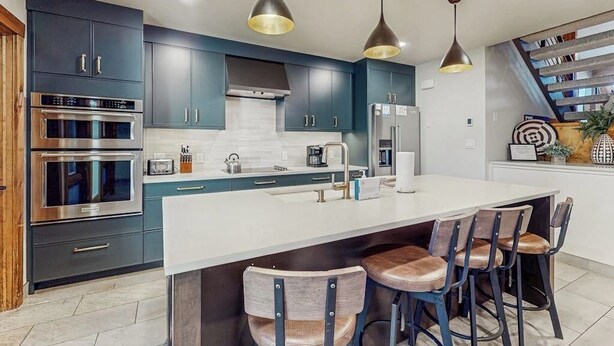 Una cocina moderna con armarios oscuros, electrodomésticos de acero inoxidable, una isla central con taburetes, iluminación colgante y una escalera con libros.