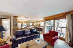 A living room with a black leather sofa, a red armchair, and a wooden coffee table. There is a kitchen in the background with wooden cabinets and a dining area. - Ground-floor home with high-speed WiFi, pool & hot tub - next to lift (Steamboat Springs)