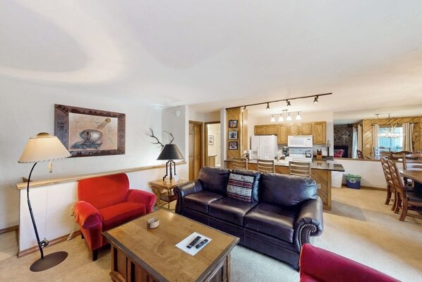 Ein Wohnzimmer mit einem roten Sofa, einem schwarzen Ledersofa, einem Couchtisch und einer Lampe. Im Hintergrund ist eine Küche mit Holzausbau und ein Essbereich mit Stühlen zu sehen.