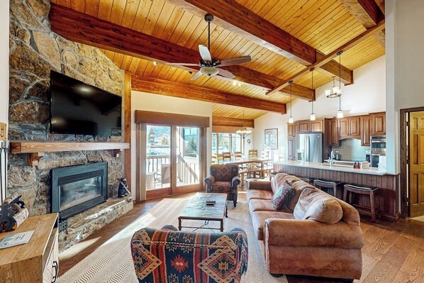 A spacious living area with a stone fireplace, a large TV, and a ceiling fan. - Luxury condo next to lift with pool, hot tubs & high-speed WiFi (Steamboat Springs)