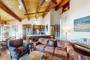 A spacious living area with a wooden ceiling, a sofa, a patterned armchair, a coffee table, and a dining area with a wooden table and chairs. - Luxury condo next to lift with pool, hot tubs & high-speed WiFi (Steamboat Springs)