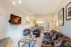 Ein modernes Wohnzimmer mit Flachbildfernseher, zwei Sesseln, einer Couch und einem Couchtisch, im Hintergrund befindet sich eine Küche.