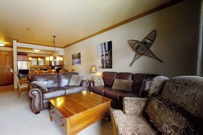 A living room with a brown leather sofa, a wooden coffee table, and patterned armchairs. There is a kitchen in the background with wooden cabinets and a dining area. - Charming condo just steps to base area w/shared pool, hot tub, free WiFi (Steamboat Springs)