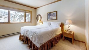 A bedroom with a large bed, a nightstand with a lamp, a framed picture on the wall, and a window with a view of trees. - Charming condo just steps to base area w/shared pool, hot tub, free WiFi (Steamboat Springs)