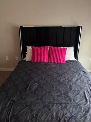Ein Bett mit schwarzem Kopfteil, einer dunkel gemusterten Bettdecke und zwei leuchtend pinkfarbenen Kissen.