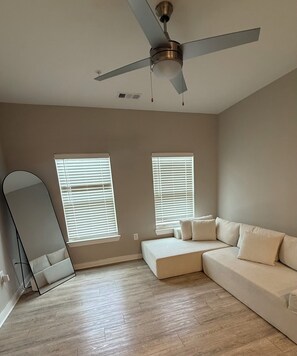 Ein Zimmer mit Deckenventilator, zwei mit Jalousien versehenen Fenstern, einem großen Spiegel und einem beigefarbenen Ecksofa.