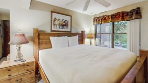 Ein Schlafzimmer mit Bett, Nachttisch, Lampe, Deckenventilator, Fenster mit Vorhängen und einem gerahmten Bild an der Wand.