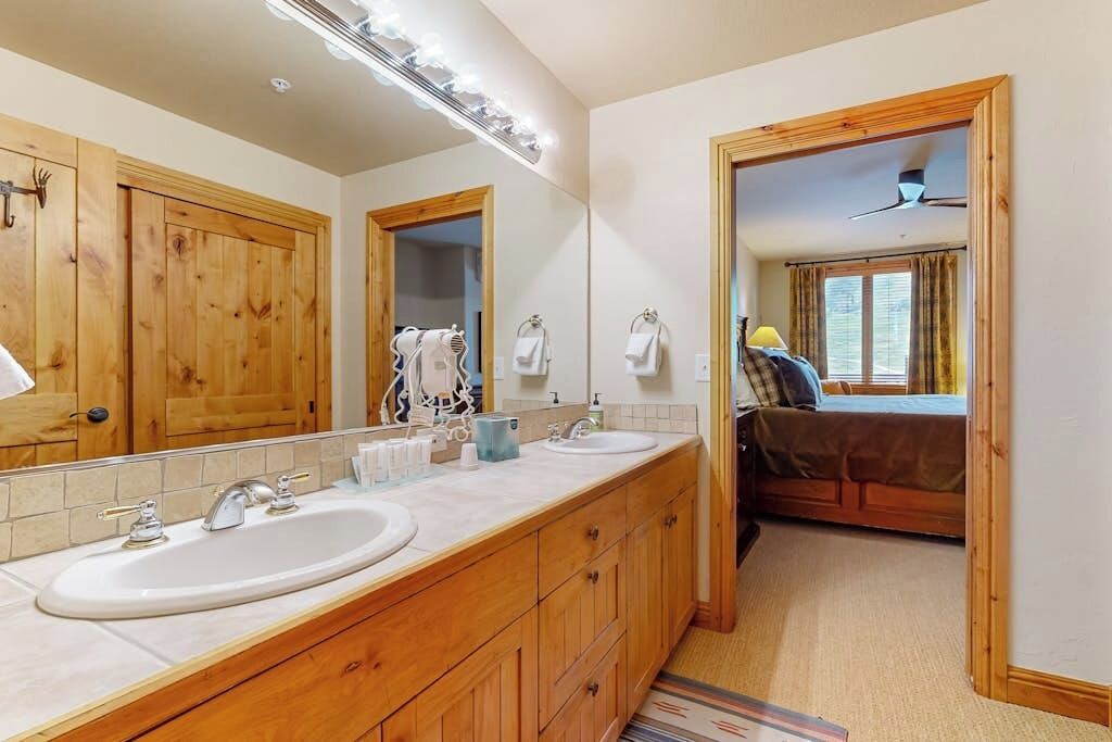 A bathroom with wooden cabinets, a large mirror, two sinks, and a bed visible through an open doorway.