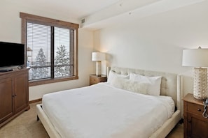 A bedroom with a large bed, a television on a wooden cabinet, a window with blinds, and bedside lamps. - Luxurious corner condo w/ view & shared pool, hot tubs, game room & more (Steamboat Springs)