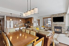 A modern kitchen with wooden cabinets, a dining area with a wooden table and chairs, a living room with a fireplace, and a mounted TV. - Luxurious corner condo w/ view & shared pool, hot tubs, game room & more (Steamboat Springs)