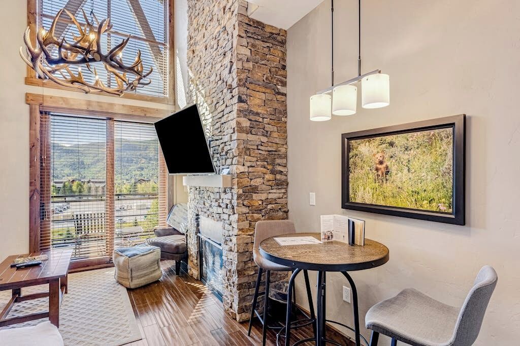 A modern living room with a stone fireplace, a mounted TV, a round table with chairs, and a large window offering a view of the outdoors.