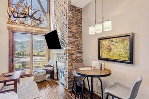 Ein modernes Wohnzimmer mit Kamin aus Stein, einem an der Wand montierten Fernseher, einem runden Tisch mit Stühlen und einem großen Fenster mit Blick ins Freie.