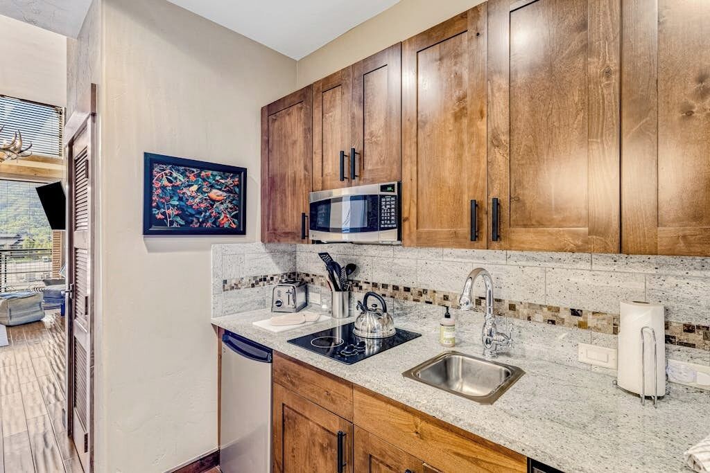 Una cocina con armarios de madera, microondas, lavabo y encimera con tetera y utensilios.