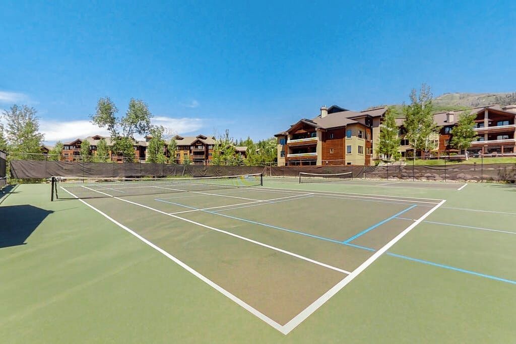 Una cancha de tenis rodeada de edificios con balcones, cielo despejado y árboles.
