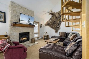 A living room with a stone fireplace, leather sofas, a mounted television, and a fur wall piece. - Loft condo with Flattop Mountain views, shared hot tubs, gas fireplace, & gym (Steamboat Springs)