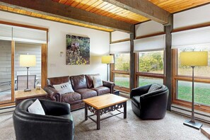 Ein Wohnzimmer mit Holzbalkendecke, großen Fenstern, einem braunen Ledersofa, einem schwarzen Sessel und einem Holztisch.
