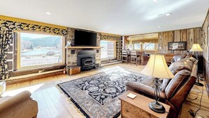 A living room with a fireplace, a sofa, a TV, and a large window overlooking a scenic landscape. - Dog-friendly riverfront retreat with hot tub, stunning views & screened porch (South Fork)