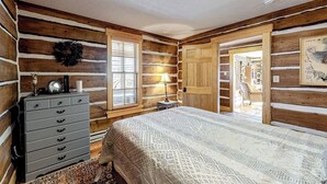 A log cabin-style bedroom with a bed, dresser, lamp, and a view of a dining area. - Dog-friendly riverfront retreat with hot tub, stunning views & screened porch (South Fork)