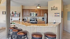 A kitchen with wooden cabinets, a granite countertop, a stainless steel refrigerator, and a bar area with stools. - The Lodge at Steamboat A301 Upscale condo with balcony, pool, hot tubs, & fireplace - walk to lifts (Steamboat Springs)